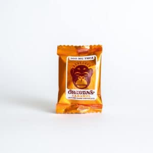 A small dark chocolate bar called "Xite Origins Caramel Chocolate Bar - 300mg," orange-wrapped, infused with 300mg THCA, and featuring a cartoon monkey face on the packaging.