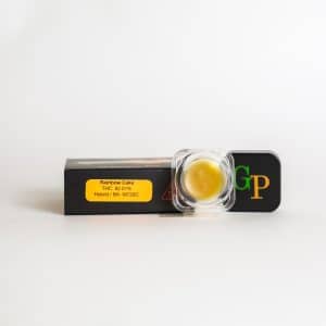 A small container of yellow cannabis concentrate sits on a black box labeled "Green Crack Sugar Wax - Label No.420 - 1g," with THC content and hybrid strain information displayed on an orange sticker.